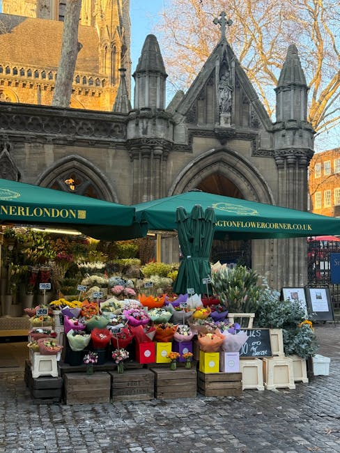 A vibrant outdoor flower stand set up in front of a historic stone building with Gothic architectural features, including pointed arches and turrets, in Stoke Newington. The display is under a large g