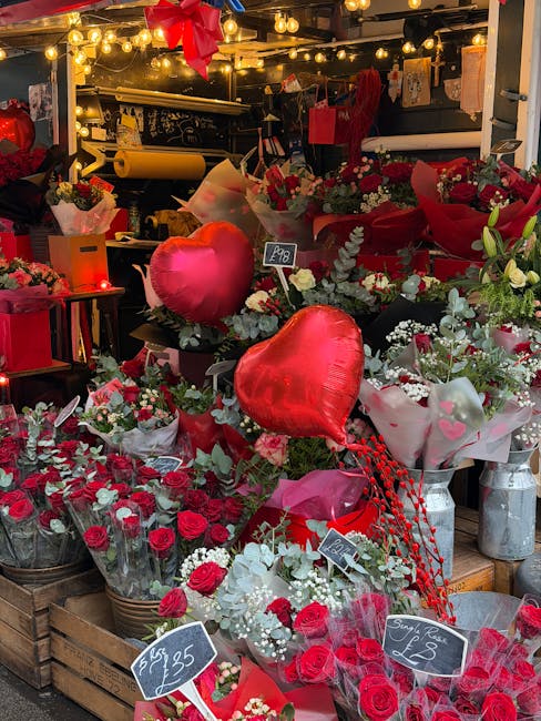 A vibrant display of floral arrangements at a flower shop, featuring red and white roses, baby's breath, and green foliage. The bouquet arrangement is wrapped in clear cellophane and pink tissue paper