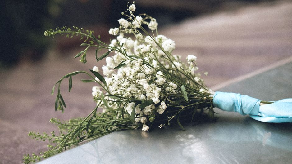 A bouquet of white Gypsophila flowers with delicate, tiny blooms and slender green stems, wrapped in a light blue satin ribbon, lying on a smooth surface with a blurred outdoor background. The arrange