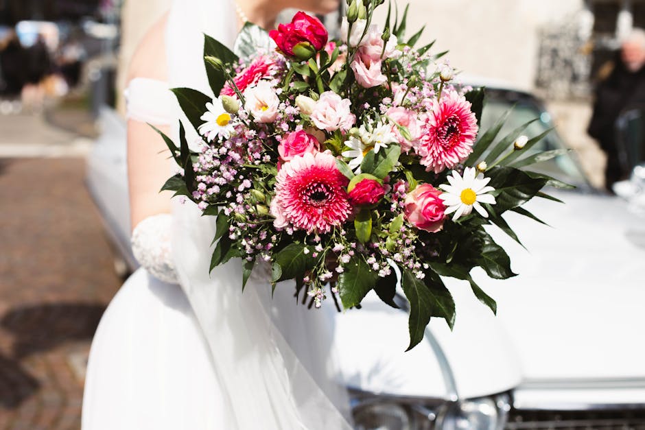 A bride holds a vibrant floral bouquet featuring pink and white gerbera daisies, pink roses, white daisies with yellow centers, and delicate pink baby's breath, arranged with lush green foliage. The b