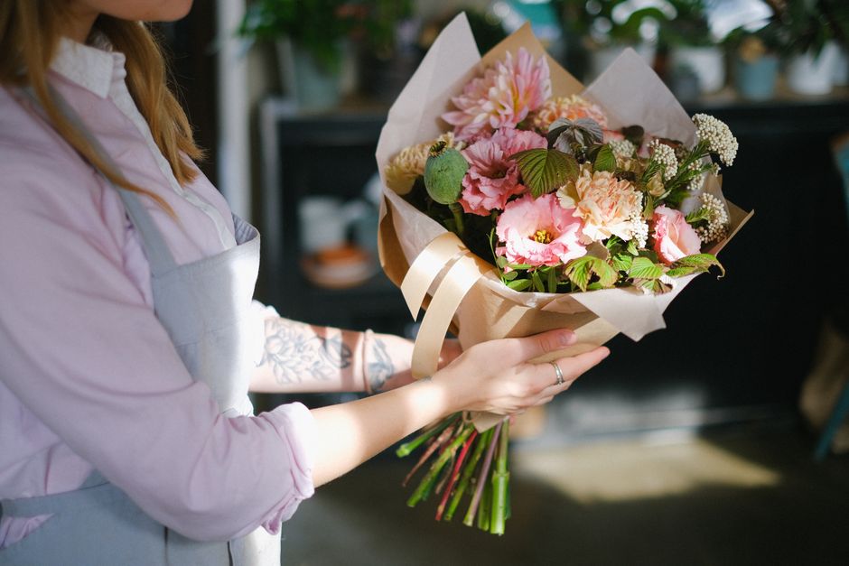 A person holding a freshly arranged floral bouquet featuring pink and cream flowers, including roses, carnations, and filler blooms, wrapped in beige tissue paper secured with a cream ribbon. The bouq